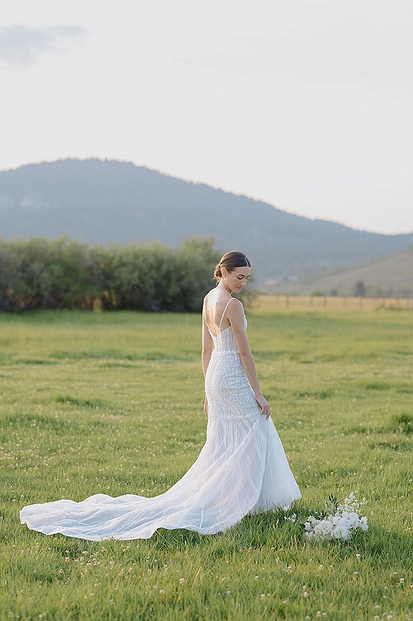 Bridal portrait of a bride in a backless spaghetti-strap gown with long train, looking over shoulder in a meadow with mountains behind