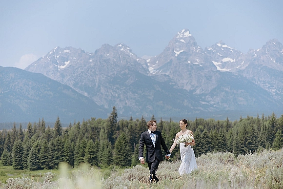 Couple portrait of newlyweds holding hands, bride with a simple white bouquet and groom in a tuxedo before mountain meadows and pines