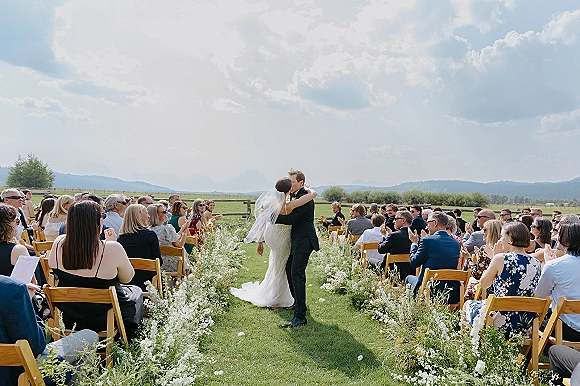 Ceremony kiss during an outdoor wedding ceremony, bride in flowing veil and gown with groom in suit on a floral-lined aisle in a mountain meadow