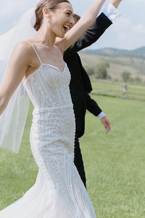 Wedding recessional with bride raising her hands in veil and dress beside groom in black tuxedo on a grassy lawn with hills and sky
