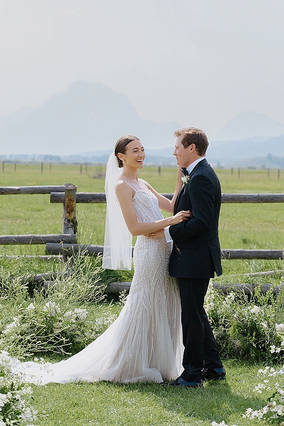 Couple portrait of bride and groom laughing as she touches his face, veil and tuxedo in a mountain meadow by a wooden fence