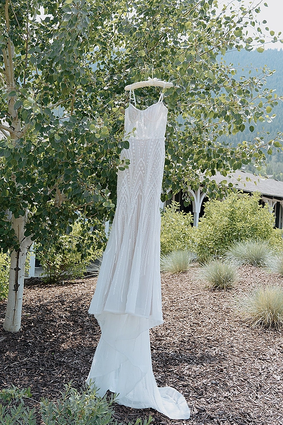 Wedding dress in lace hanging on a hanger with spaghetti straps, framed by leafy branches with mountains and a building behind