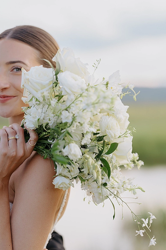 Bridal portrait of a bride holding bouquet of white roses covering her face, gold ring stack visible against grassy hills and sky