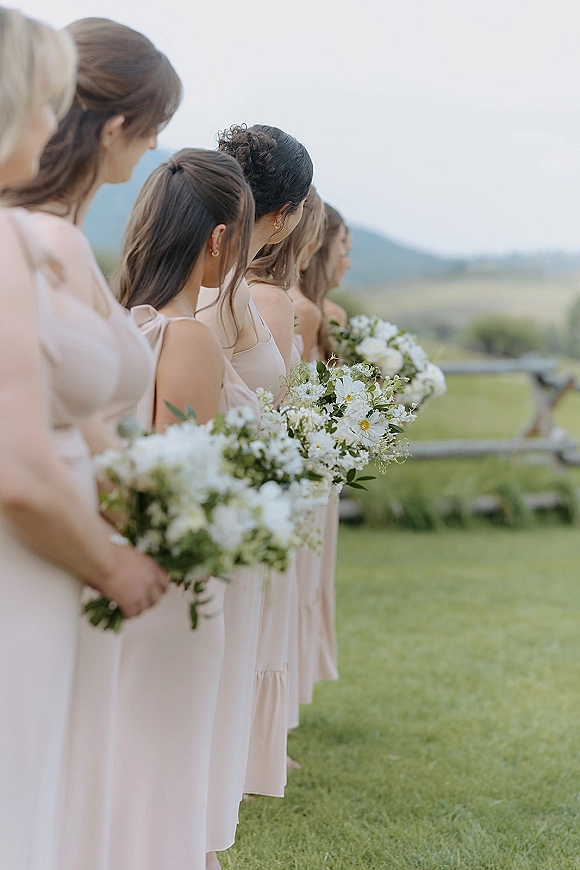 Bridesmaid lineup in blush dresses holding white and green bouquets on an outdoor lawn by a wooden fence with rolling hills beyond
