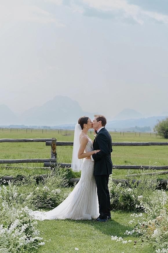 Wedding kiss portrait of bride and groom kissing, her veil flowing beside a split rail fence in a meadow with distant mountains under clouds