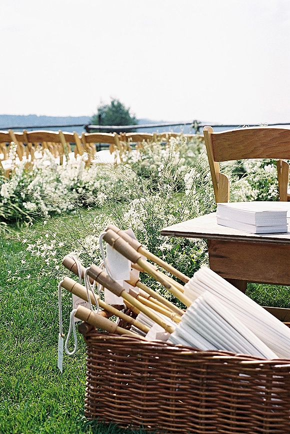 Ceremony aisle decor with outdoor ceremony chairs lined with wildflowers, wicker fan basket and programs on a grassy lawn with hills beyond