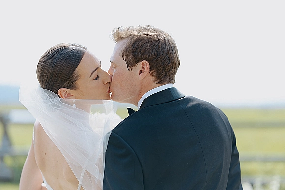 Wedding kiss portrait of bride and groom kissing in side profile, veil blowing, groom in black tux and bow tie by waterfront field