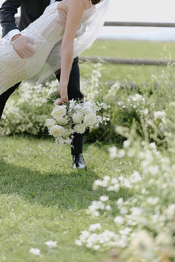 Wedding couple portrait of groom lifting bride into a dipped kiss, her veil and white rose bouquet flowing in a wildflower field by a fence