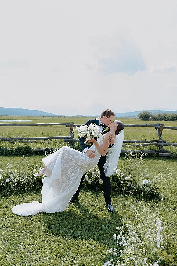 Wedding kiss portrait of groom dipping bride as she holds a bouquet, veil trailing over grass with rustic fence and mountains behind