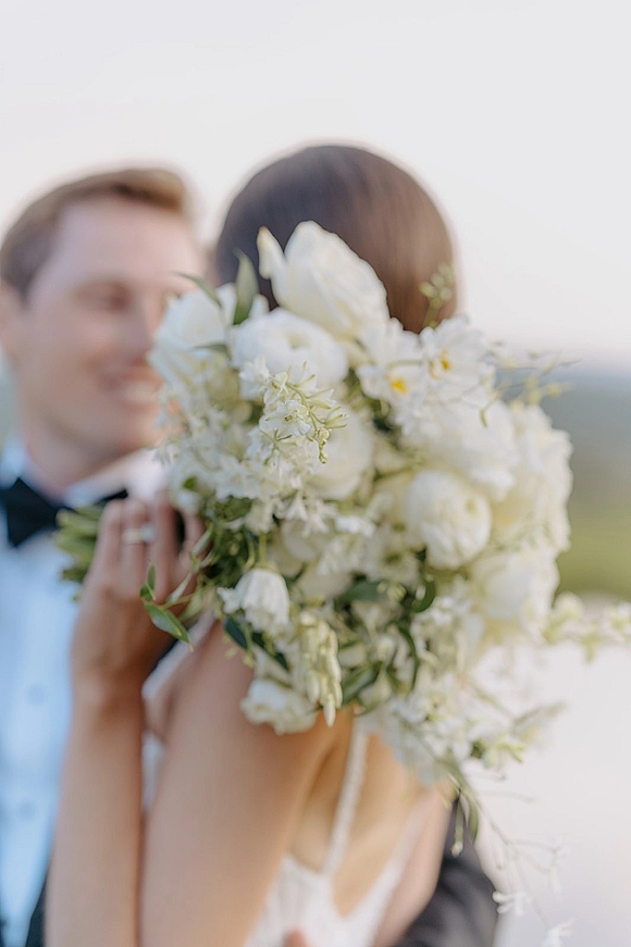 Couple portrait of bride holding bouquet, bouquet covering face as groom embraces her in tuxedo and bow tie under open sky