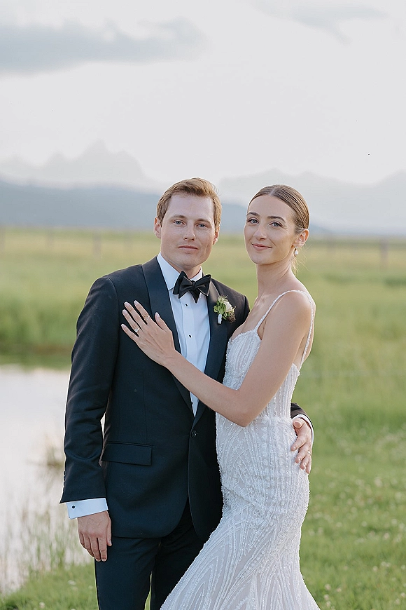 Couple portrait of bride in sleeveless wedding dress and groom in black tuxedo embracing in meadow by pond with mountains under clouds