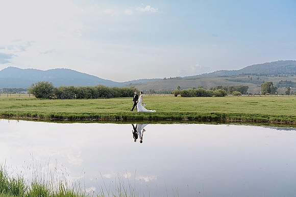 Couple portrait of bride and groom walking by a reflective pond, bride holding bouquet with veil and long train, mountains behind under clouds