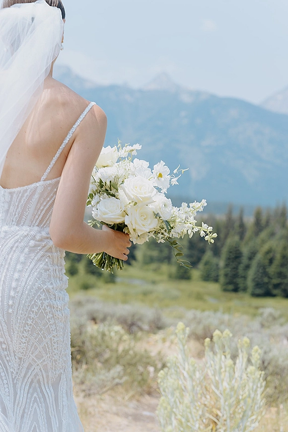 Bridal portrait of a bride holding bouquet, seen from behind with a long veil in an alpine meadow with evergreens and mountains