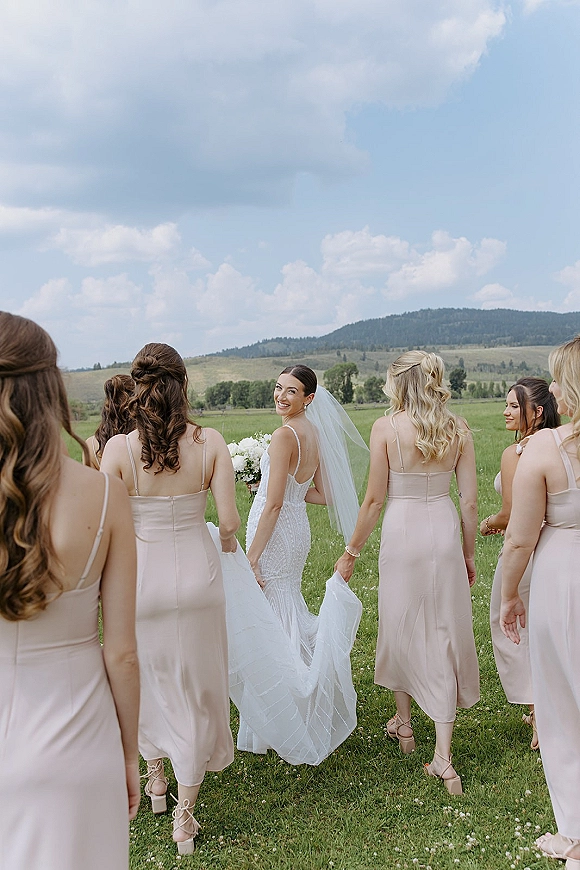 Bridal party photo of bride with bridesmaids walking in a meadow, veil and bouquet flowing, with mountains under a cloudy sky