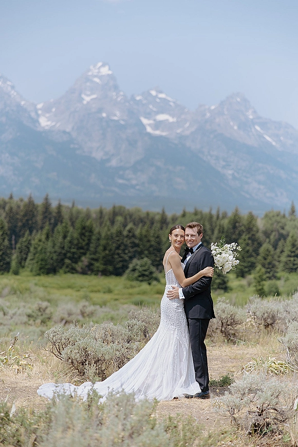 Couple portrait of bride and groom embracing, her veil and white bouquet against a mountain range and evergreen meadow backdrop