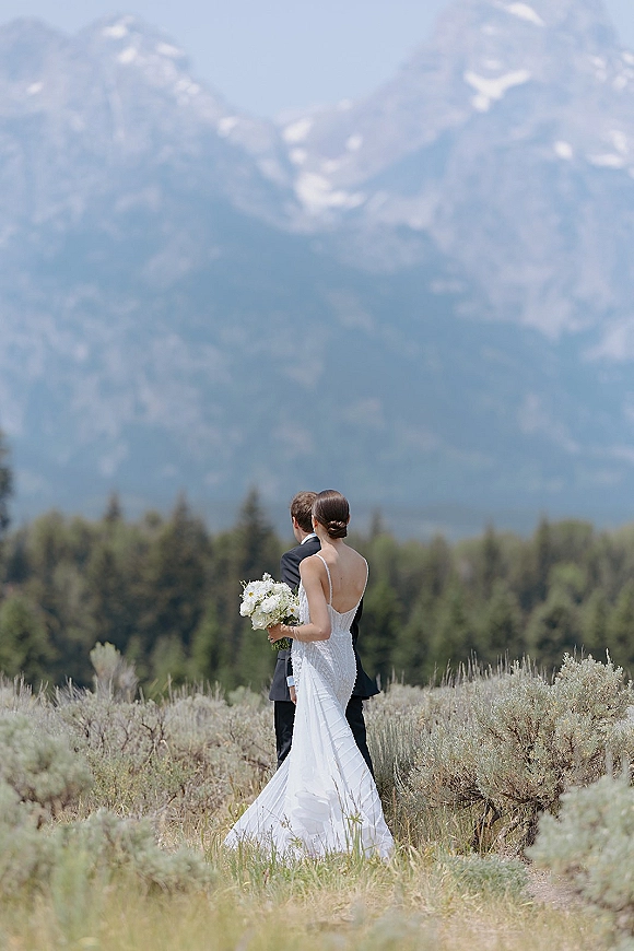 Couple portrait from behind, bride and groom holding a bouquet in a meadow with sagebrush, evergreens, and distant mountains under blue sky