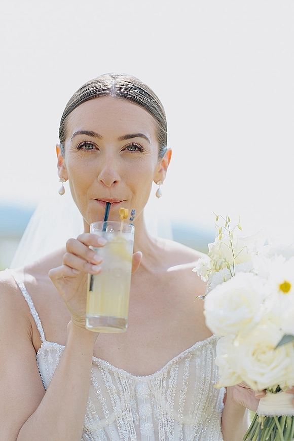 Bridal portrait of a bride holding bouquet and sipping a cocktail through a straw, wearing a veil and pearl drop earrings under a bright sky