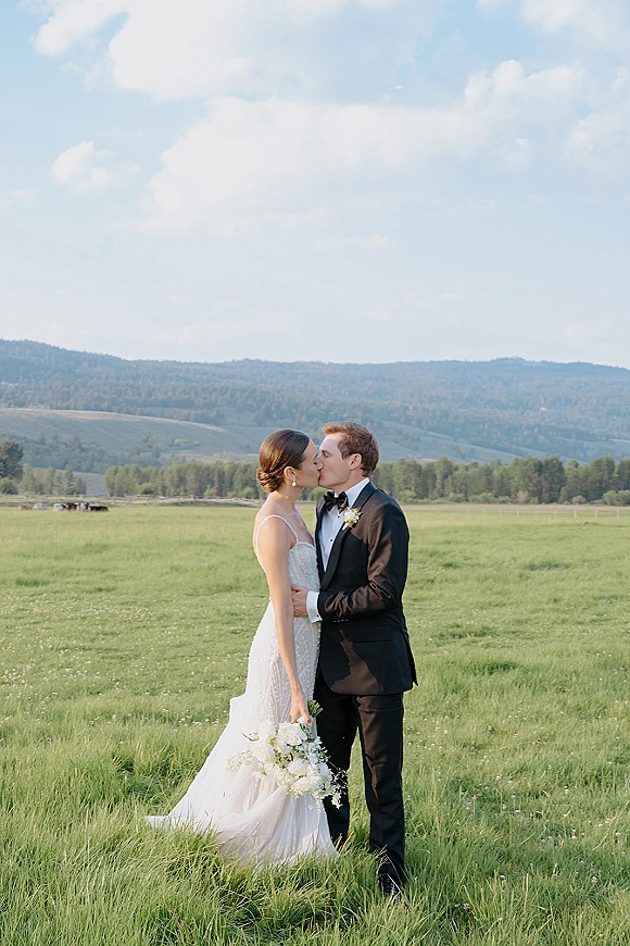 Wedding kiss portrait of bride and groom kissing in a grassy meadow, bride holding a white bouquet with mountains beneath blue sky clouds