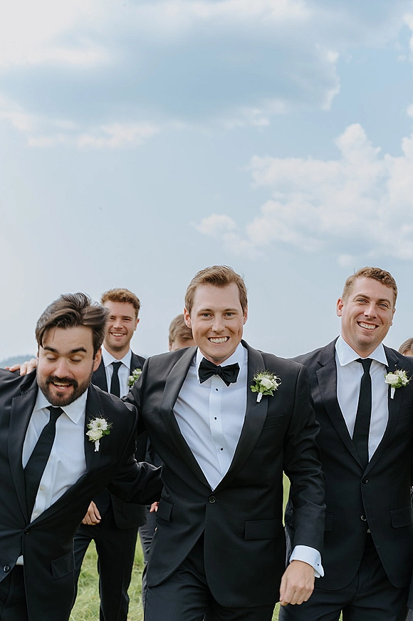 Groomsmen portrait of the groom with groomsmen in black tuxedos with boutonnieres, walking across a grassy field under a blue sky