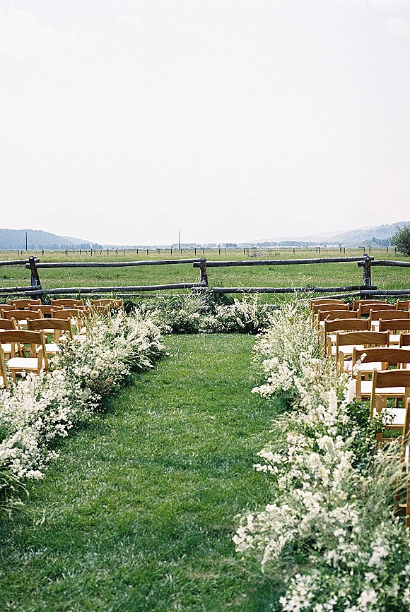 Ceremony aisle decor with white florals and greenery lining an outdoor wedding aisle, framed by wood folding chairs in an open field