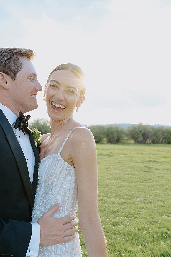 Couple portrait of bride laughing in a beaded wedding dress and groom in tuxedo with bow tie, embracing in a sunlit field with trees