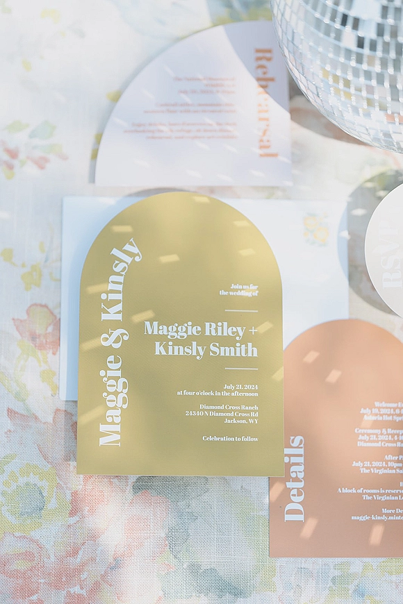 Wedding invitations with arched cards, RSVP and details card in bold printed typography, styled on floral fabric with a disco ball and soft shadows
