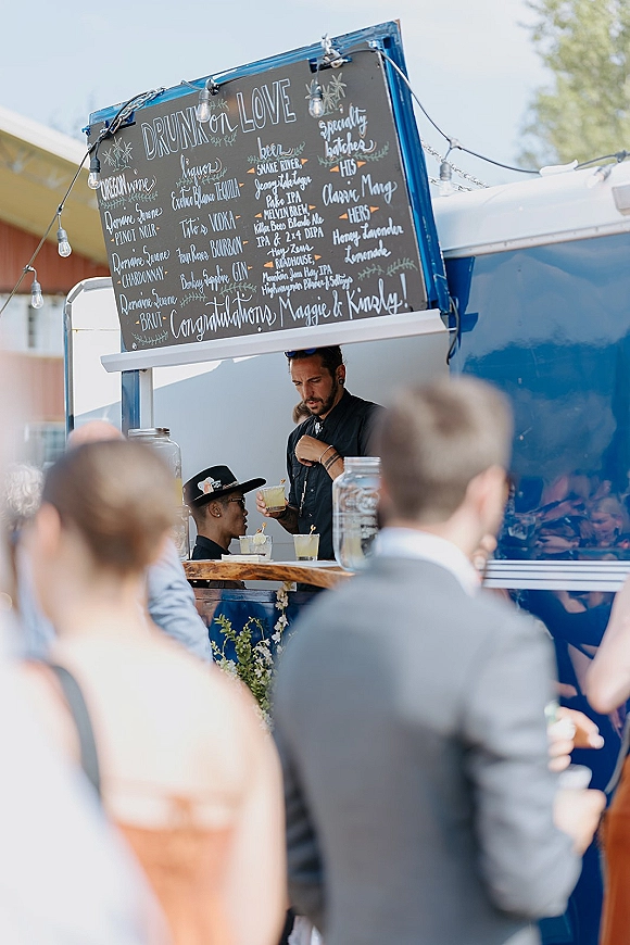 Wedding bar truck with a chalkboard drink menu, string lights, and mason jar tip jars serving cocktails to guests outdoors by a blue truck