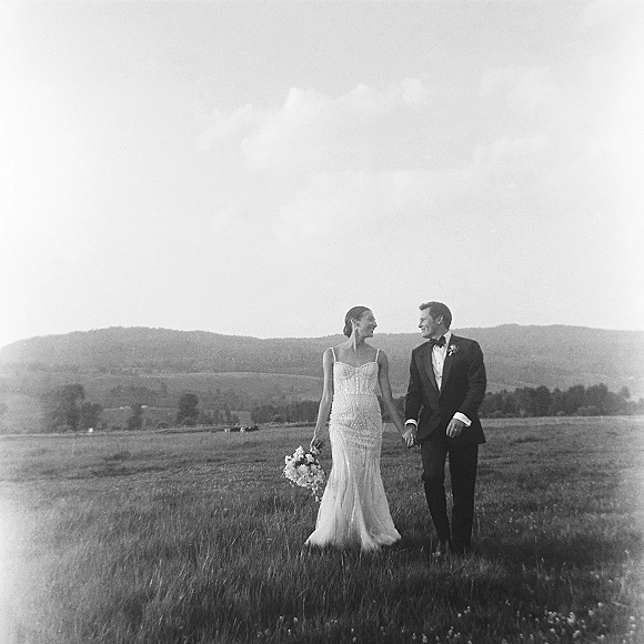 Couple portrait of bride and groom walking hand in hand, bride holding bouquet, in a meadow field with mountains under clouds