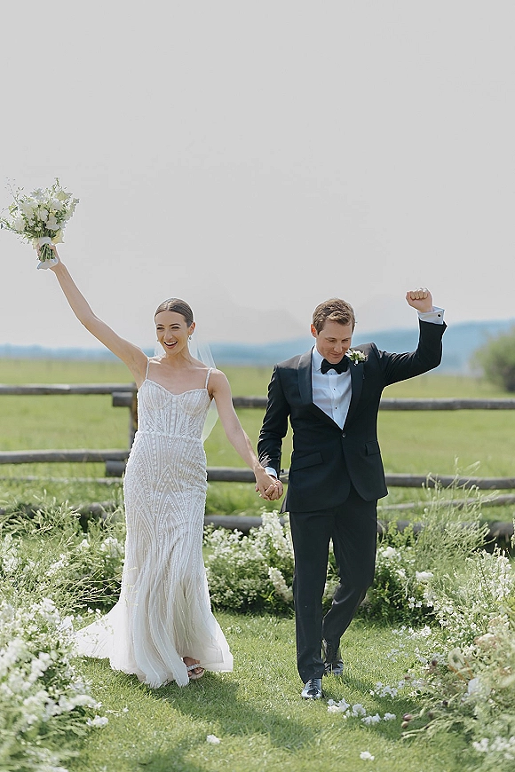 Recessional moment as bride and groom walking hand in hand, bride lifts bouquet beside groom in tux, on a wildflower lawn by a wooden fence