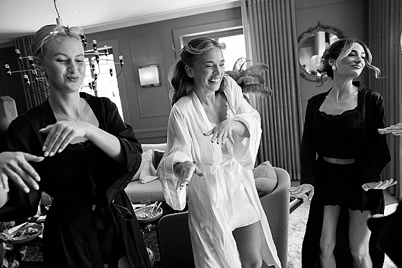 Bridal party getting ready in a hotel suite, bride and bridesmaids in robes laughing by window light with chandelier accents