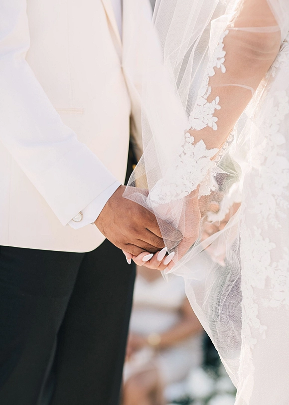 Couple holding hands in a wedding hand holding photo, showing bride’s lace sleeve, veil drape, manicured nails, and groom’s suit jacket in soft blur