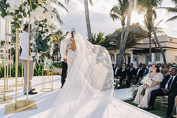 Wedding ceremony moment as bride in lace dress with cathedral veil walks to groom at white rose arch on sunny resort lawn with palms