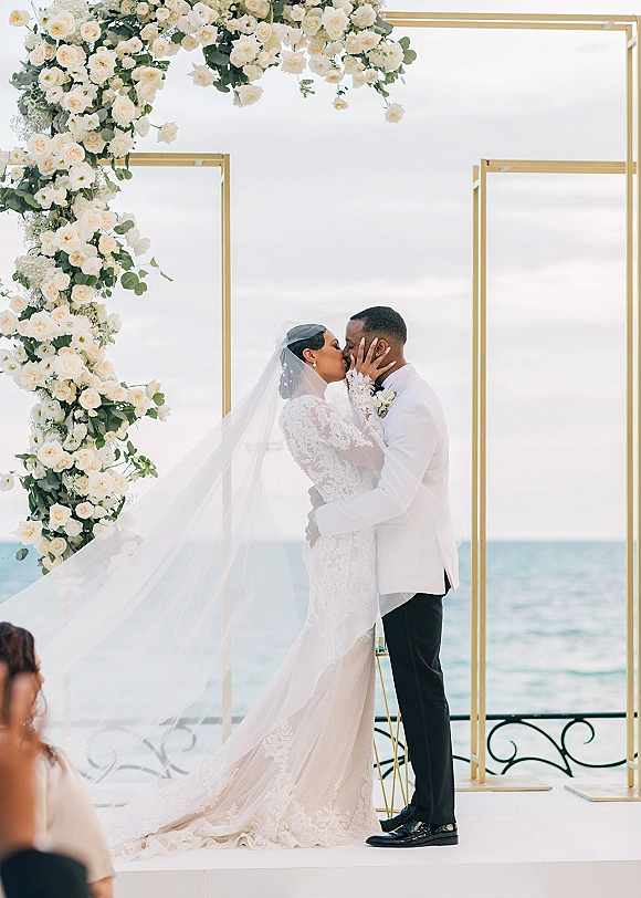 Wedding kiss beneath a white rose and greenery arch with gold frame, bride’s cathedral veil and lace dress by the ocean backdrop