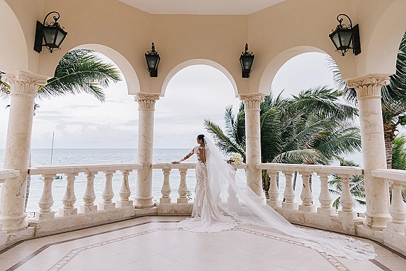 Bridal portrait of a bride with long veil in a lace wedding dress holding a white bouquet on an arched balcony overlooking ocean and palms