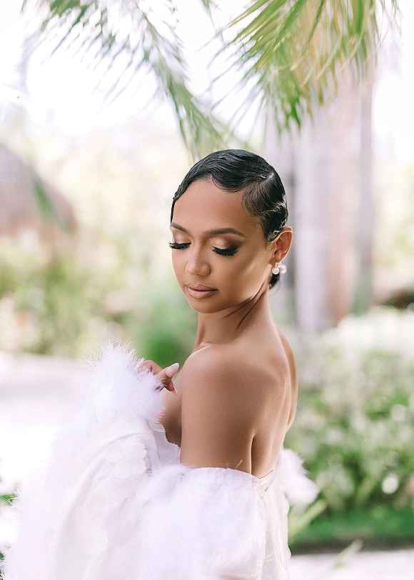 Bridal portrait in a strapless wedding dress with tulle sleeves, pearl drop earrings, and sleek finger waves against blurred palm greenery outdoors