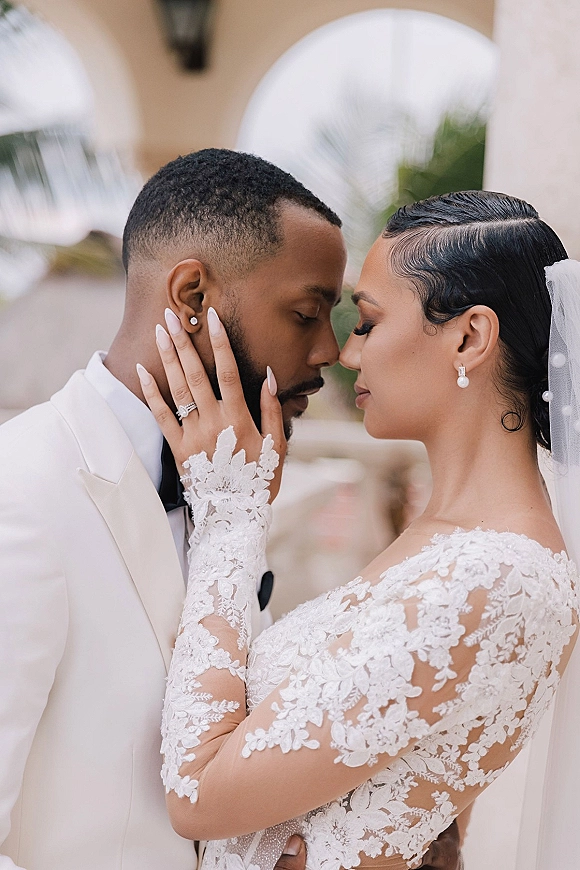 Wedding couple portrait with bride and groom close up, forehead touching as she holds his face, veil and lace gown in arched courtyard