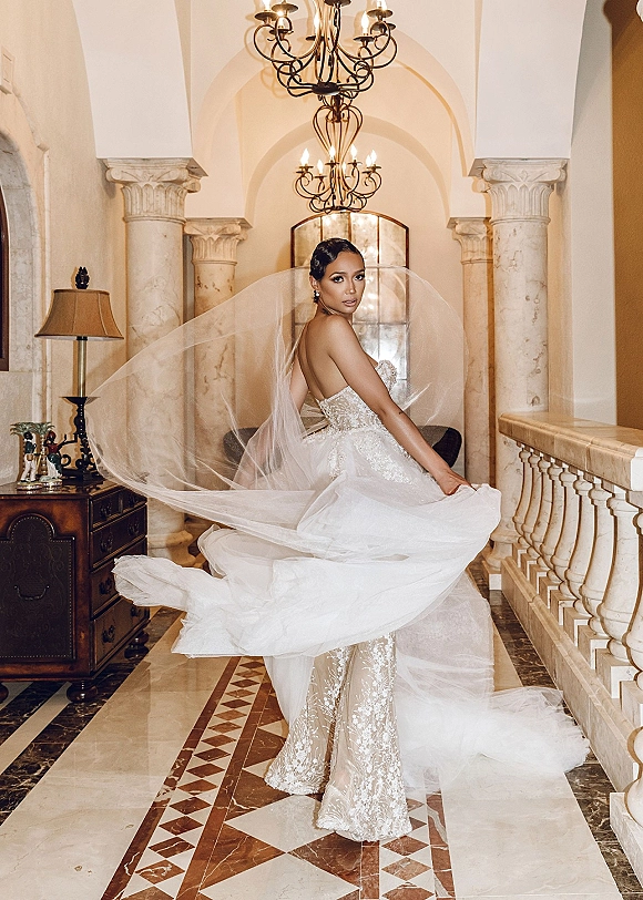 Bridal portrait of a bride in a strapless lace gown with a cathedral veil flowing behind her in an arched marble hallway with chandelier
