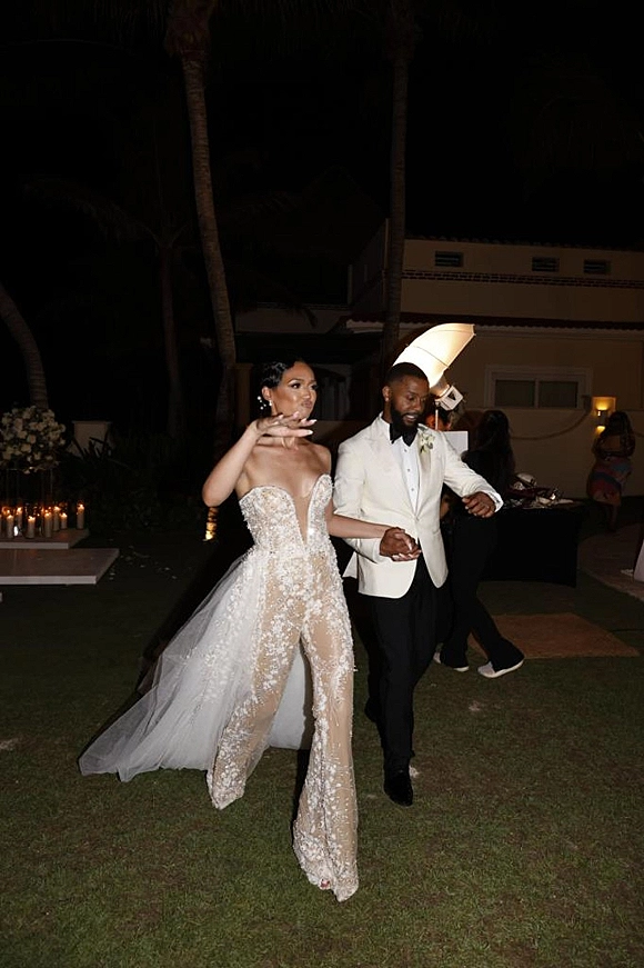 Wedding reception entrance as bride in sparkly strapless gown leads groom in white dinner jacket past candles on a palm-lined lawn at night