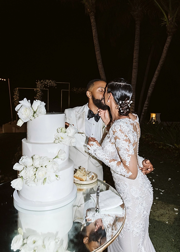 Cake cutting moment as bride and groom kiss beside a three-tier white wedding cake with roses on a glass table under string lights at night