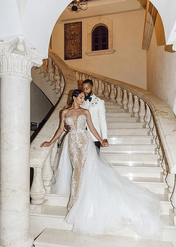 Couple portrait of bride and groom on stairs holding hands, her strapless lace gown with long train on a grand marble staircase backdrop