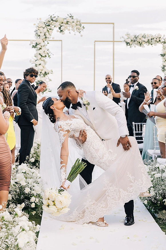 Wedding kiss moment at an outdoor ceremony recessional kiss as the groom dips the bride in a lace dress and veil beneath a floral arch
