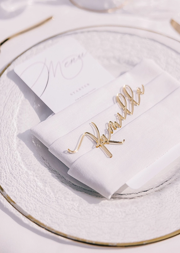 Wedding place setting with a wedding menu card atop gold rim dinner plate, folded white napkin and gold napkin ring on white tablecloth