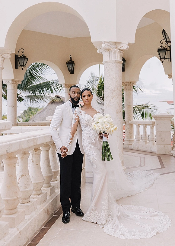 Couple portrait of bride and groom holding hands, her lace-sleeve gown and white rose bouquet under stone arches with palm trees beyond
