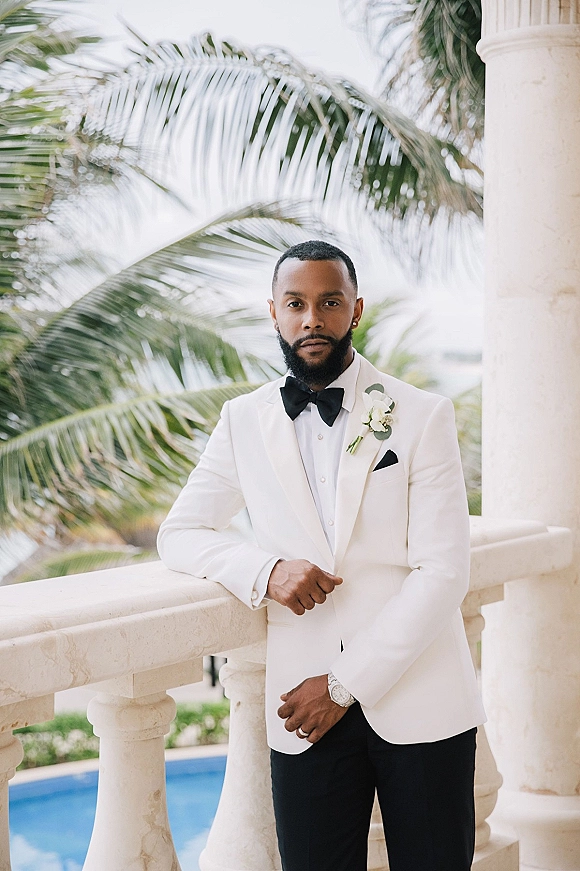 Groom portrait in a white tuxedo groom look with black bow tie, leaning on a stone balcony railing with palm trees and pool behind him