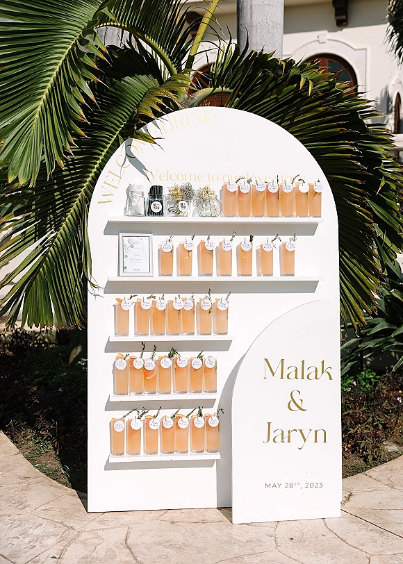Wedding welcome drinks on a welcome drink station with acrylic cups, rosemary sprigs, and round tags on an arched white sign wall outdoors