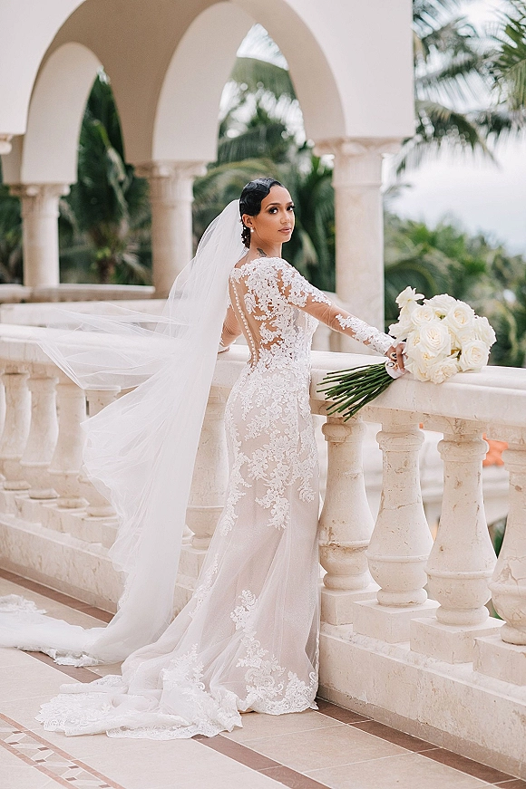 Bridal portrait of a bride in a lace wedding dress with long veil, holding a white rose bouquet on a stone terrace with palm trees