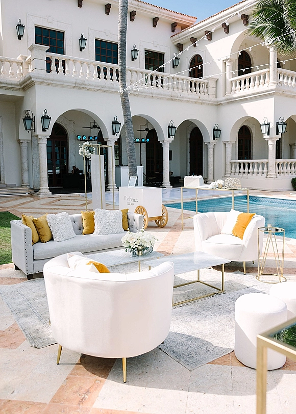 Wedding lounge setup with an outdoor wedding lounge of white chairs, gray sofa, gold pillows, and floral centerpiece beside a pool at a villa courtyard