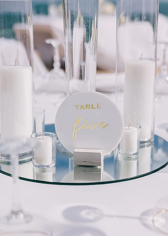 Wedding table number on a round table number sign with gold lettering, surrounded by pillar and votive candles in glass cylinders on a mirrored base