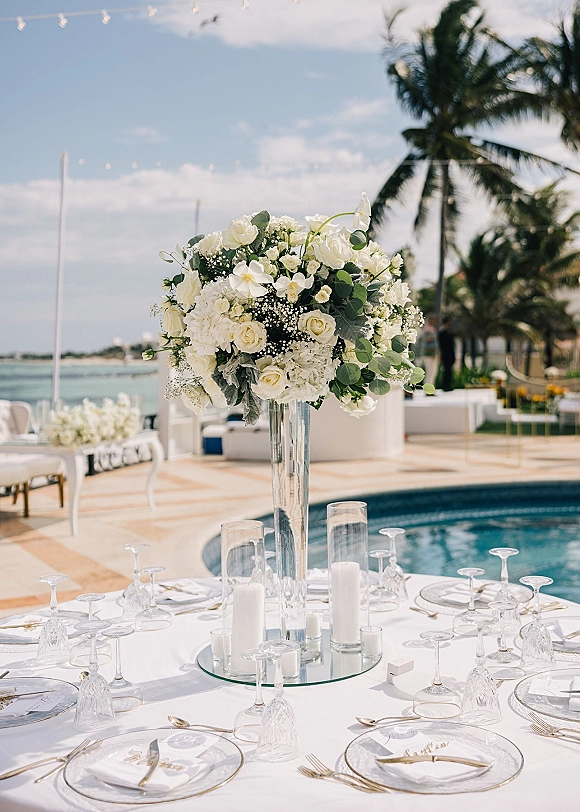 Reception tablescape with a white floral centerpiece in a glass vase, candle cylinders, gold flatware, string lights by the pool and ocean backdrop