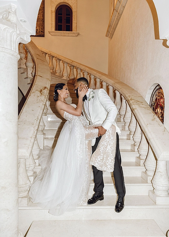 Wedding couple portrait of groom in a white tuxedo lifting his bride in a strapless lace gown on a grand marble staircase with arches